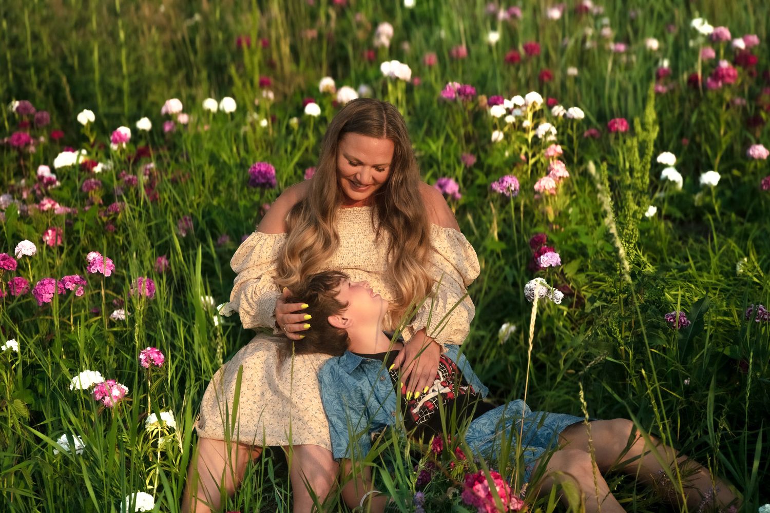 Couple sitting in a vibrant flower field at golden hour, woman in white dress smiling warmly at her partner.