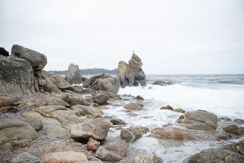 Waves crash along a rugged rocky coastline with large weathered boulders and tide pools under an overcast gray sky.