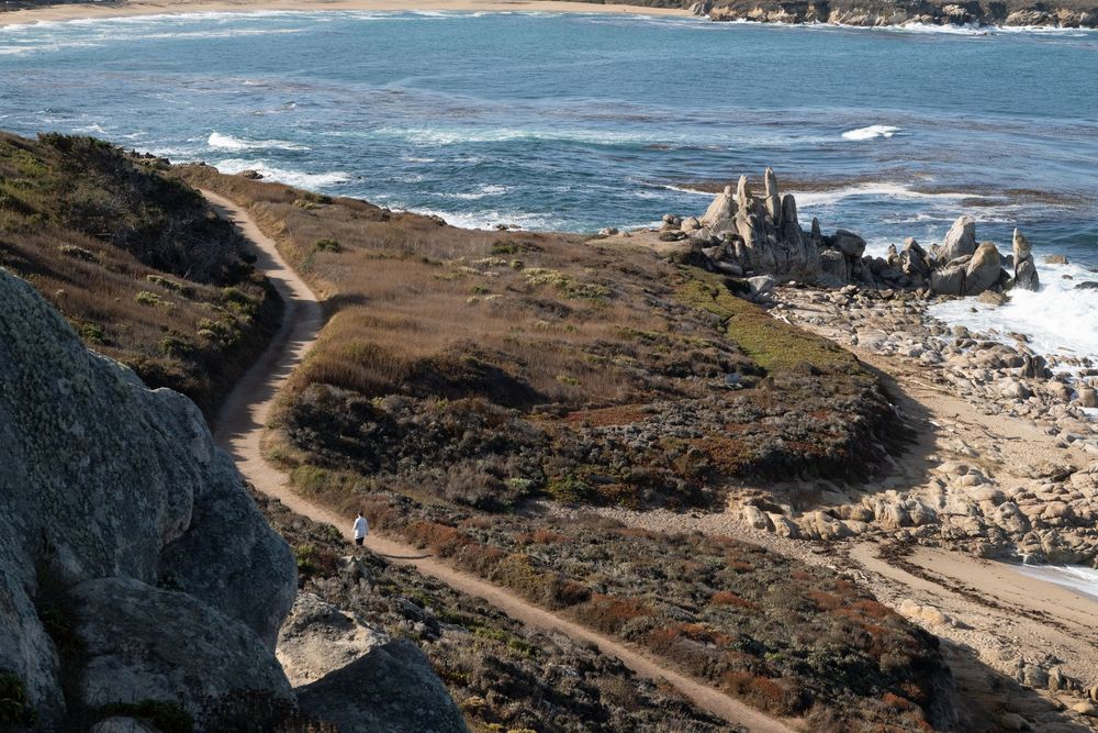 Winding dirt trail along a rugged granite coastline with rocky outcrops, coastal scrub, and turquoise sea waves crashing in the distance.