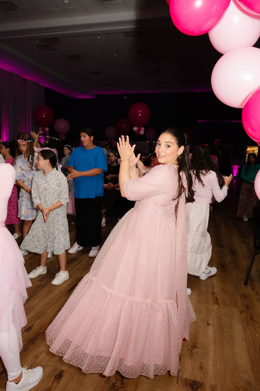 Girl in a flowing pink tulle dress dances at a party with pink balloons and guests in the background.