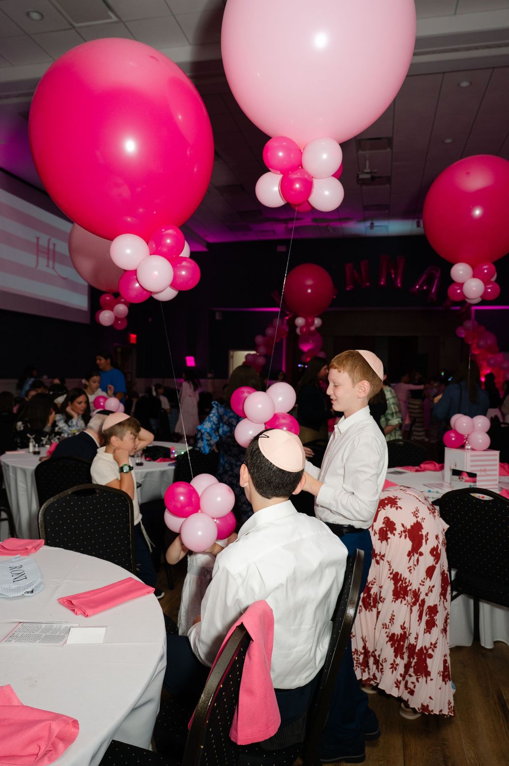 Two boys in kippahs and white shirts sit at a banquet table at a celebration decorated with pink and white balloons and pink napkins.
