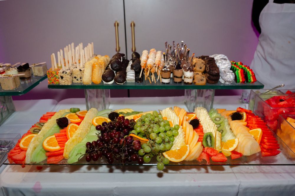 Catered buffet table with a fresh fruit display of sliced melon, oranges, grapes and berries beside an assortment of desserts and pastries.