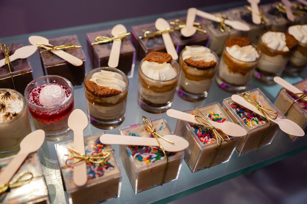 Assorted mini dessert cups with creamy layers, berry compote, and sprinkle-filled treat boxes with wooden spoons on a glass buffet display.