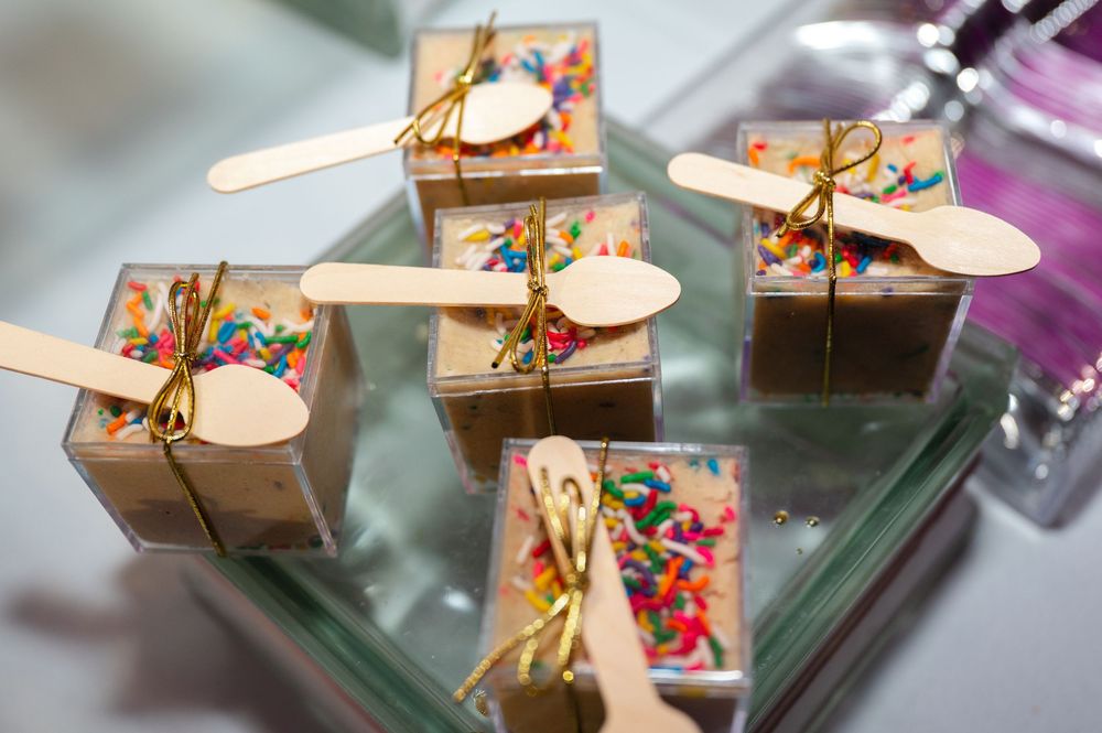 Clear dessert cups topped with colorful rainbow sprinkles, tied with gold twine and wooden spoons, displayed on a mirrored tray at a party.