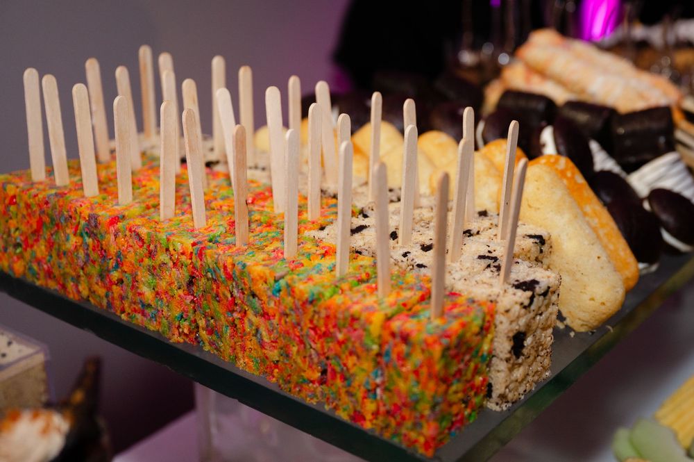 Colorful fruity cereal treats and cookies and cream bars topped with wooden sticks displayed on a glass dessert table.
