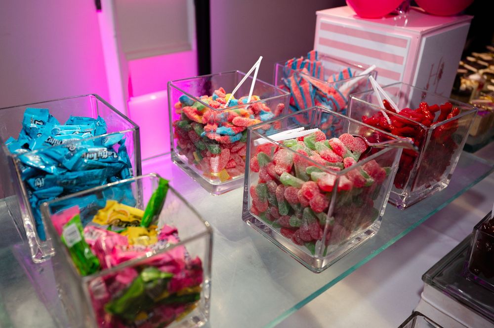Glass cubes filled with colorful candy including blue wrapped sweets, sour gummy strips and red gummies on a glass shelf at a candy buffet.