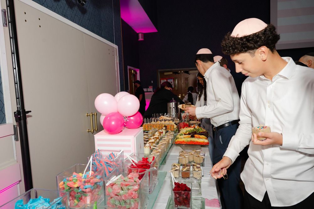 Guests in kippot browse a colorful candy and dessert buffet decorated with pink and white balloons at a festive celebration.