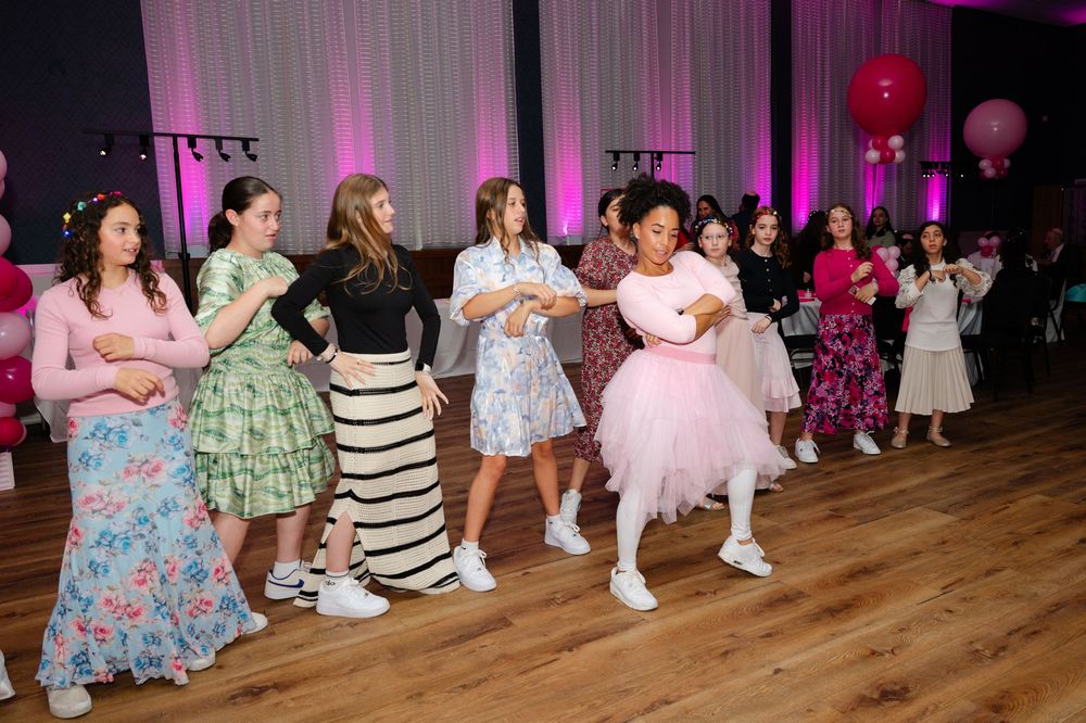 Group of girls in colorful dresses dancing together on a wooden dance floor at a festive party with pink lighting and balloons.