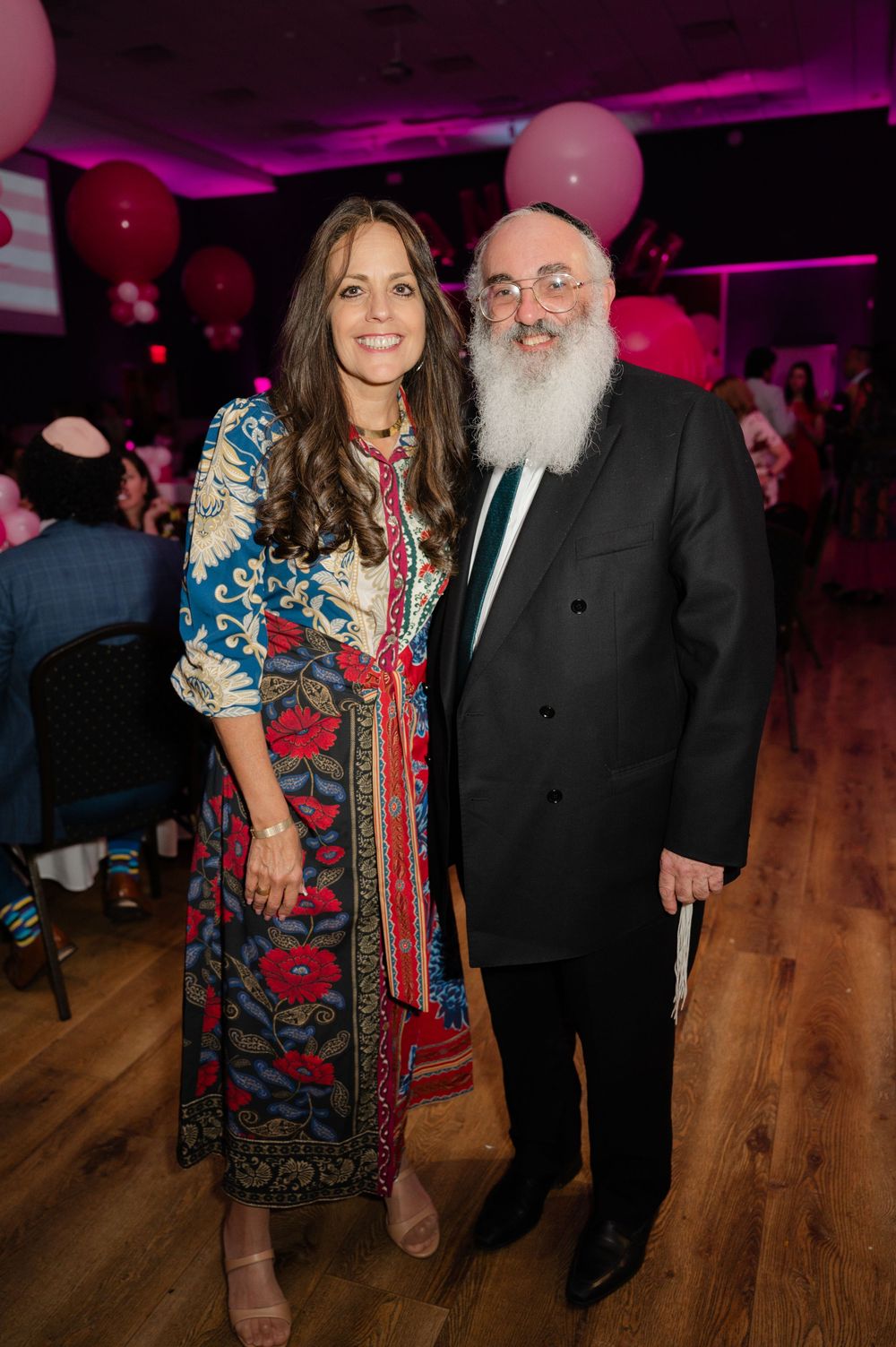 A smiling woman in a colorful floral dress poses beside a bearded man in a black suit at a pink-themed celebration.