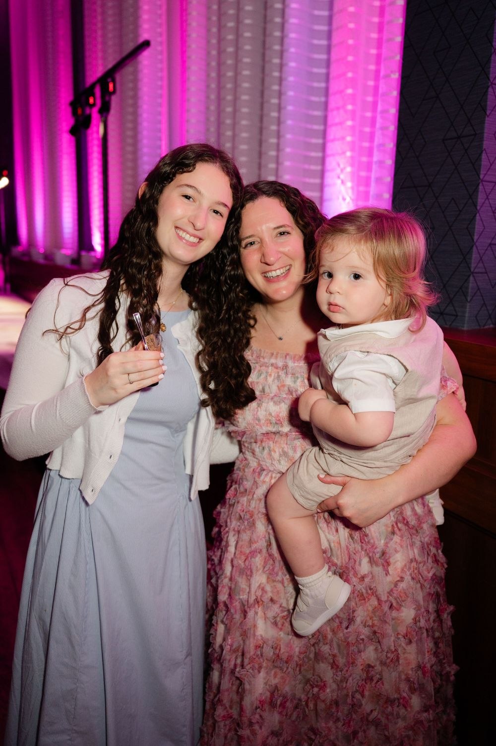 Two smiling women with long curly hair pose together at an indoor event, one holding a toddler, with pink-lit curtains behind them.