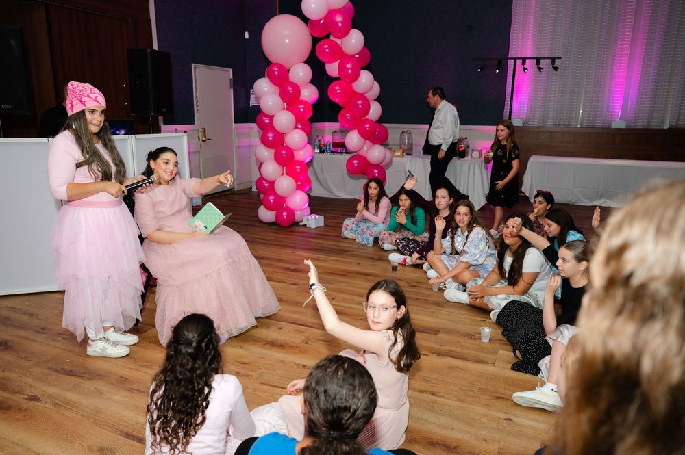 Girls in pink dresses read a story to a group of seated children at a party decorated with a pink balloon arch.
