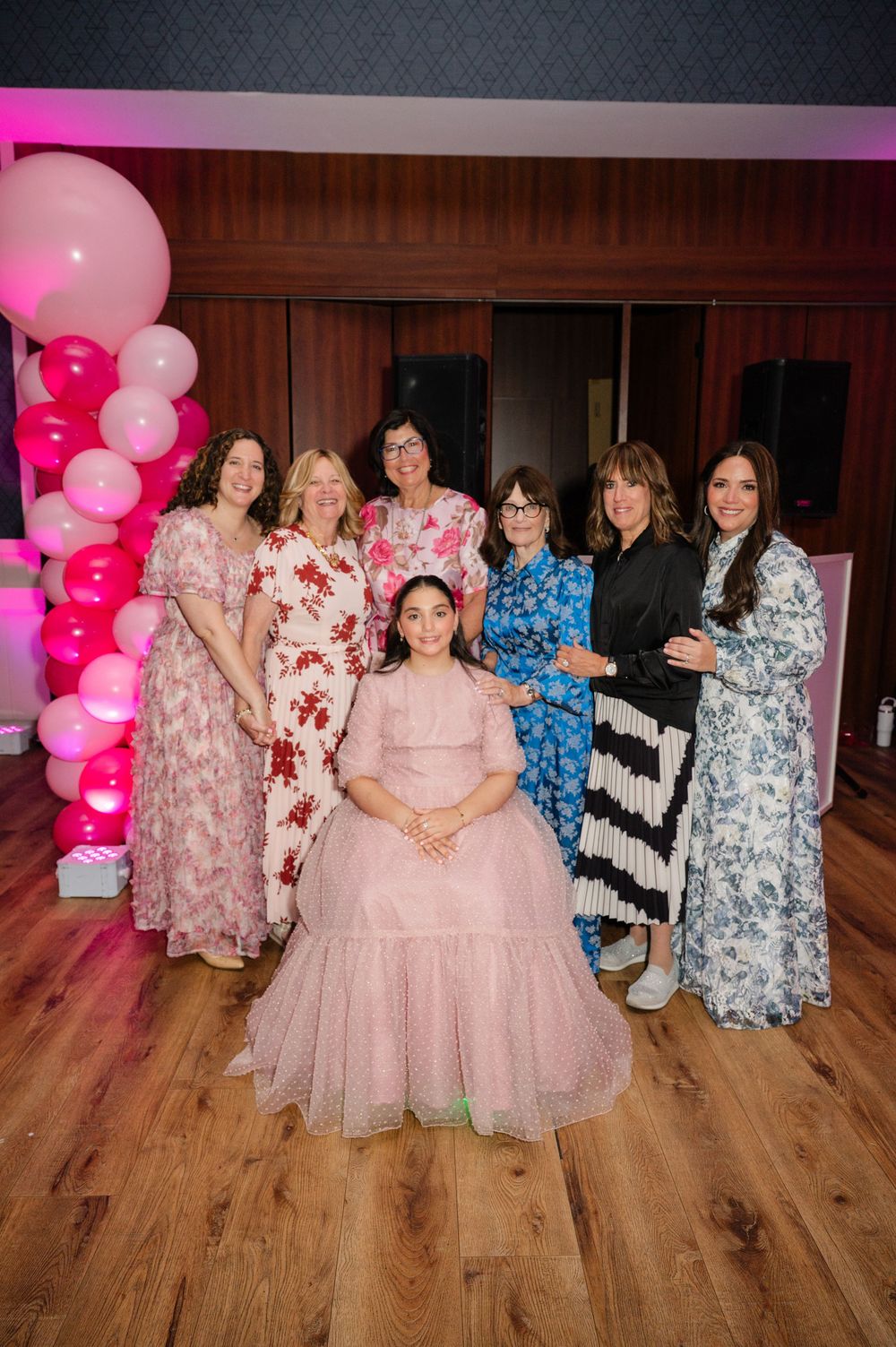 Seven women in floral and patterned dresses pose around a seated girl in a pink tulle gown beside a pink balloon garland.