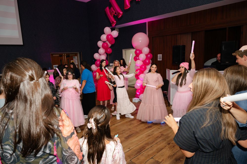 Guests in pink dresses dance and celebrate under a pink balloon garland at a festive party in a decorated hall.