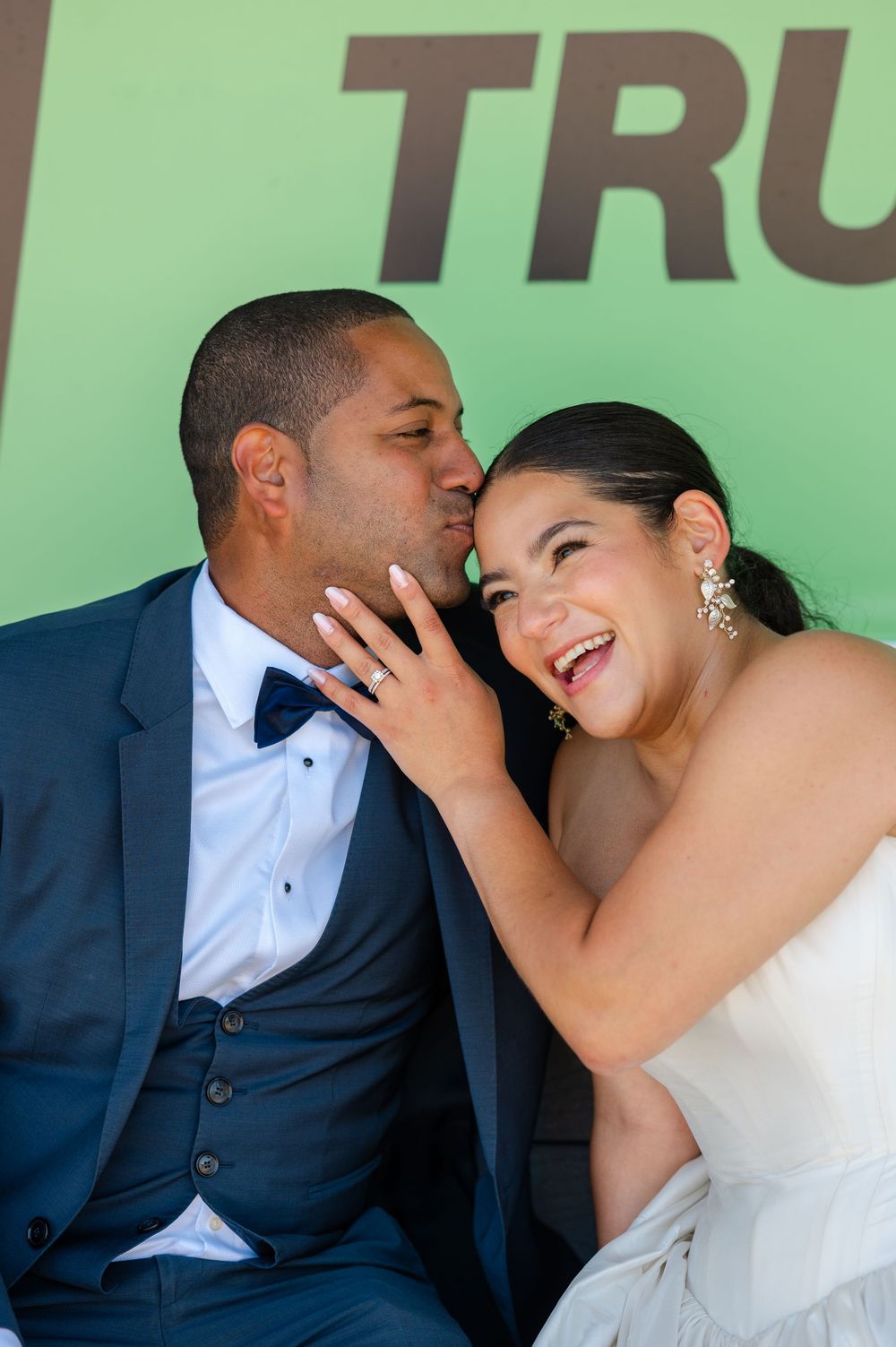 A groom in a navy suit and bow tie kisses his laughing bride in a white strapless gown on her wedding day.