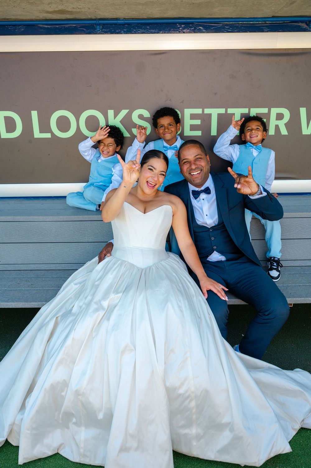 Smiling bride in a white ball gown and groom in a navy suit sit on a bench with three young boys in blue vests playfully posing.