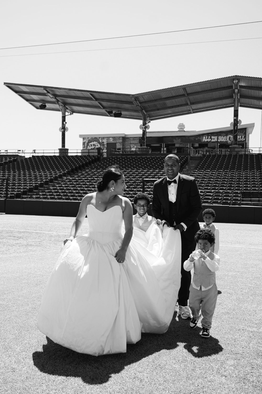 Bride in a strapless gown and groom in a tuxedo walk with two young boys on a baseball field in front of empty stadium seats.