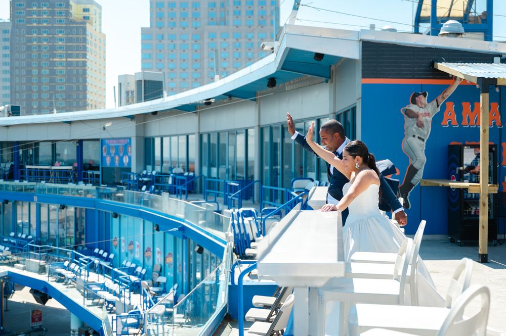 Bride in a white gown and groom in a suit embrace on a stadium terrace with blue seats, city skyline and a mascot statue behind them.