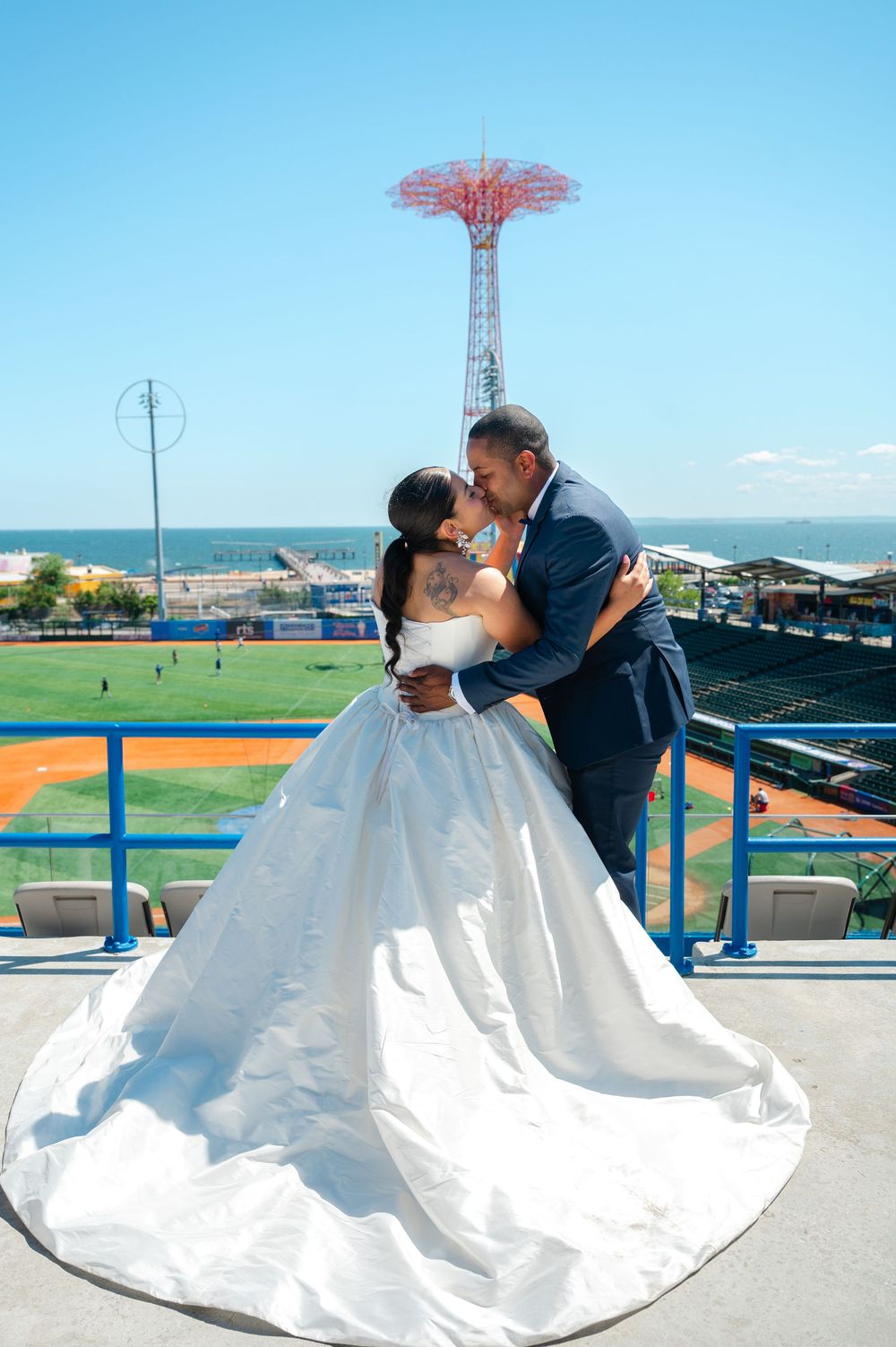 Bride in a flowing white gown and groom in a navy suit embrace on a ballpark terrace with the Parachute Jump tower and ocean behind.