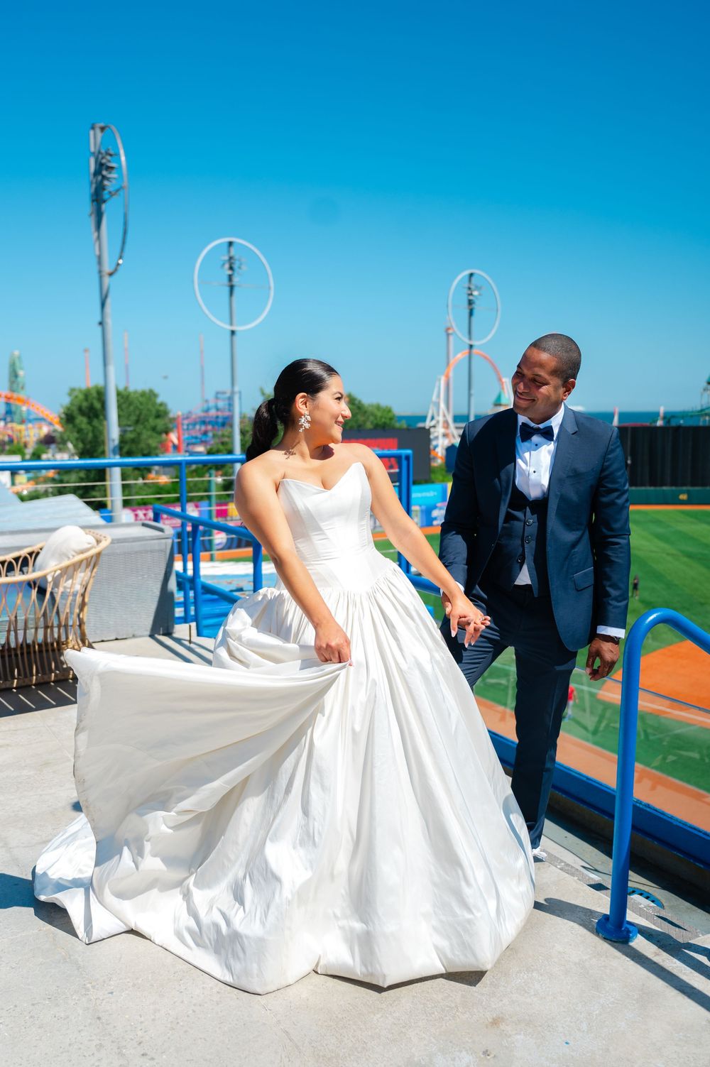 A bride in a flowing white strapless gown twirls her skirt beside a groom in a navy suit and bow tie on a sunny ballpark rooftop.