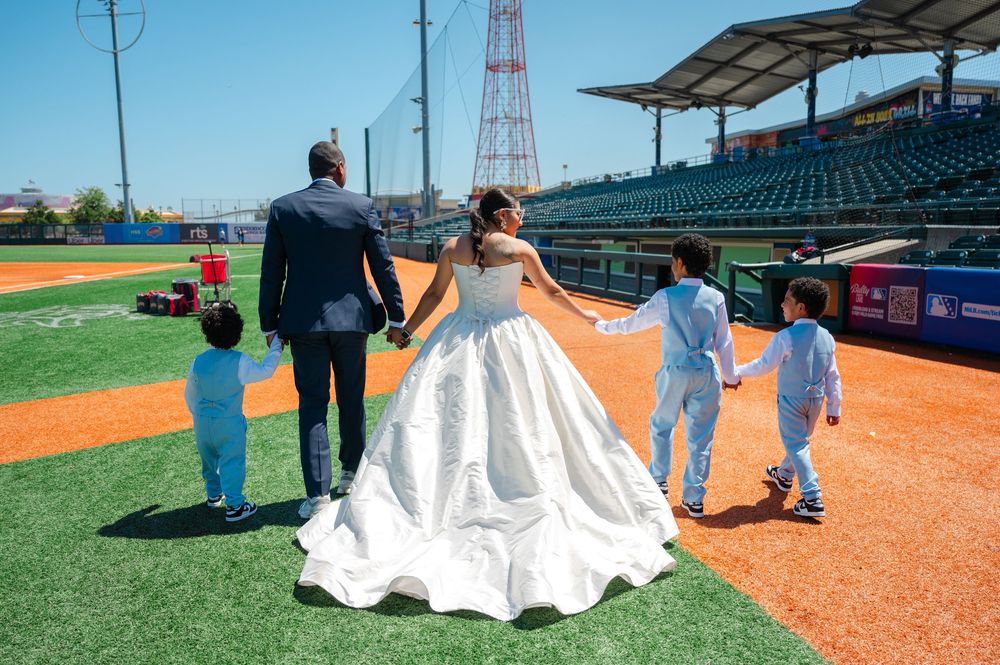 Bride in a white gown and groom in a dark suit walk hand in hand with two young boys across a baseball field during a wedding photo shoot.