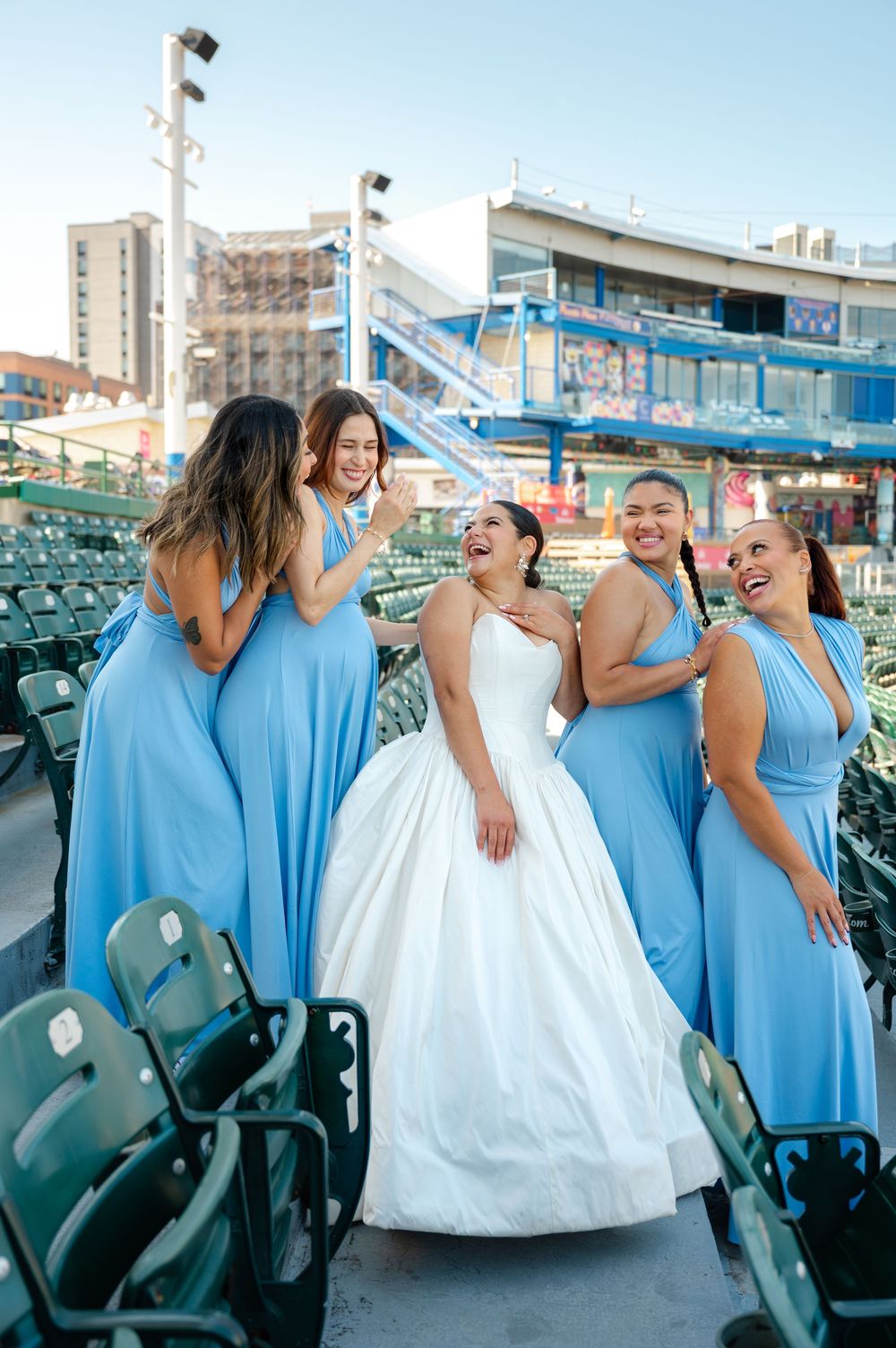 Bride in a white ball gown laughs with four bridesmaids in light blue dresses among stadium seats at a ballpark wedding.