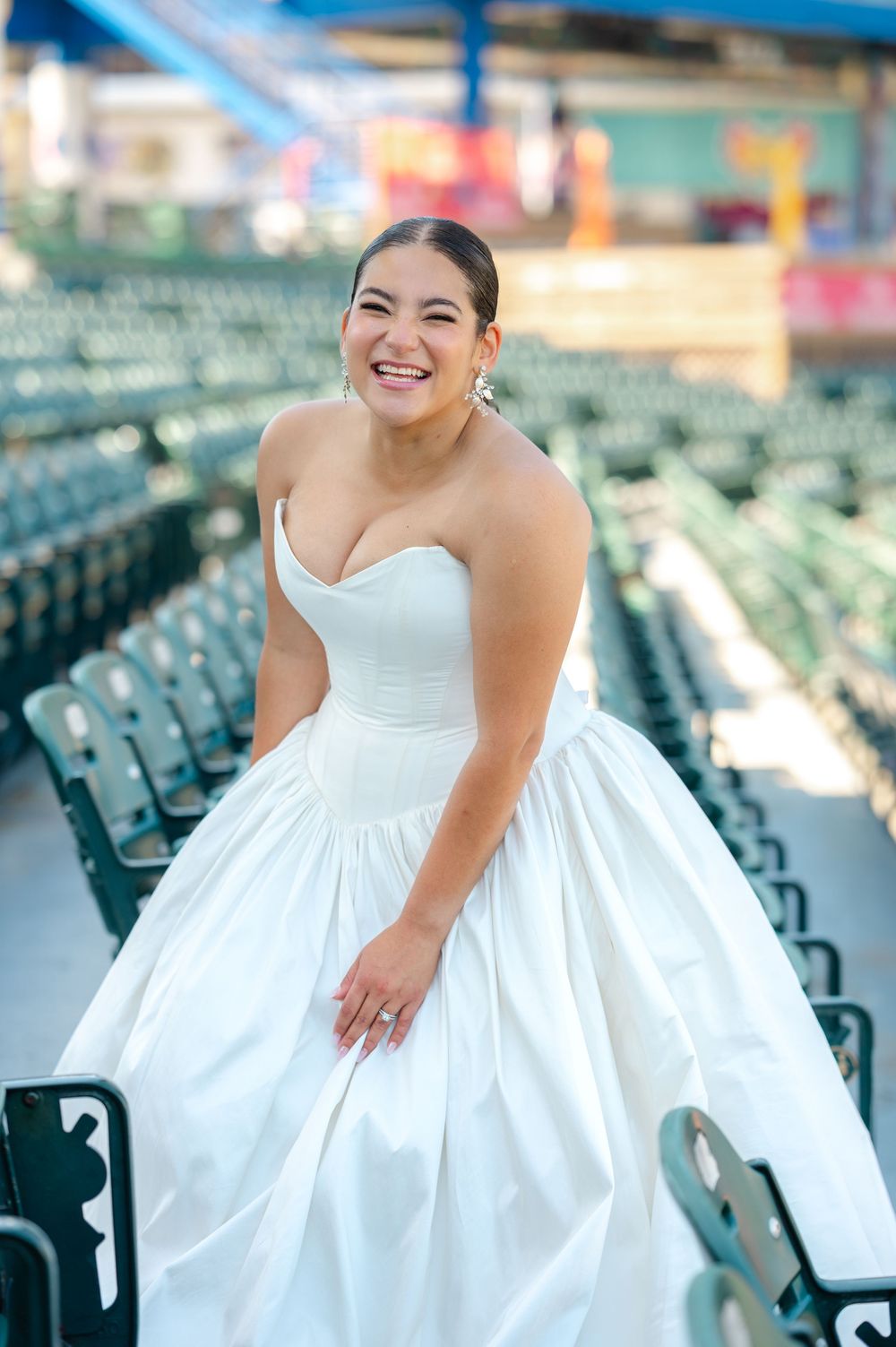 Smiling bride in a strapless white ball gown poses among empty green stadium seats at a ballpark wedding portrait session.