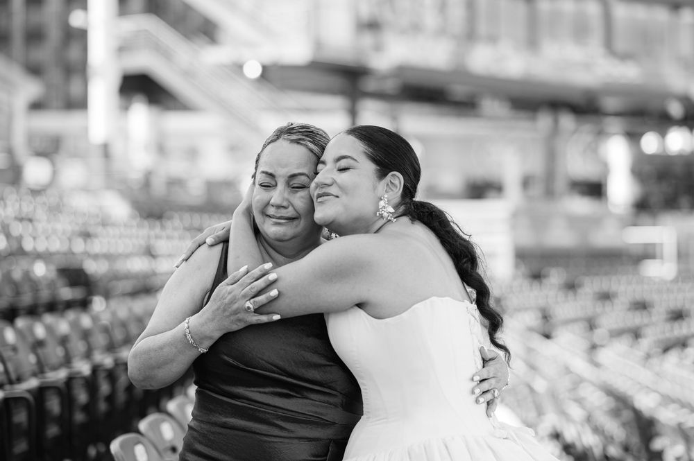 Black and white photo of a bride in a white gown embracing an older woman in a dark dress in an empty stadium during a wedding.