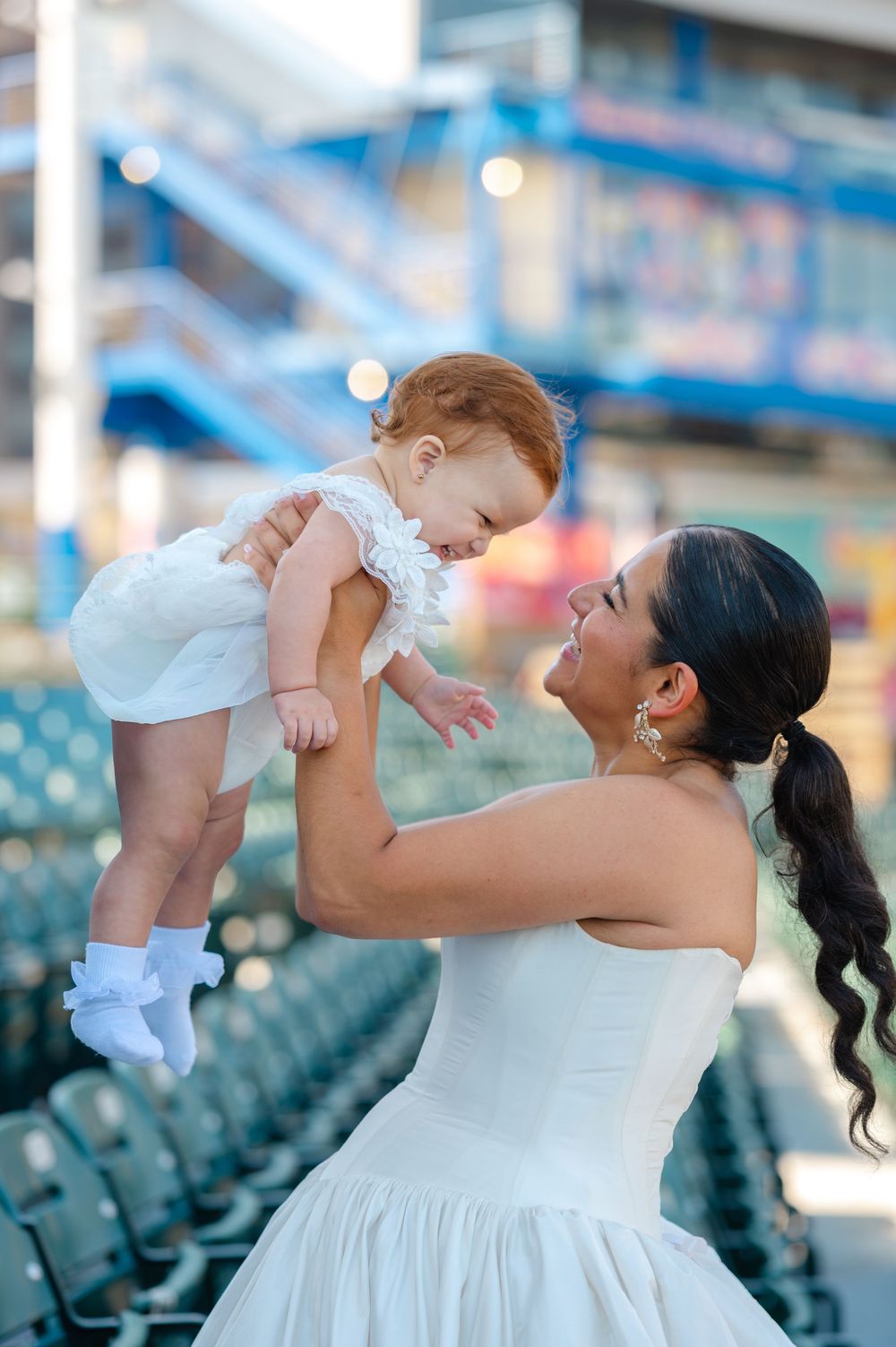 Smiling woman in a white dress lifts a giggling baby in a white outfit above her in a stadium with empty green seats.