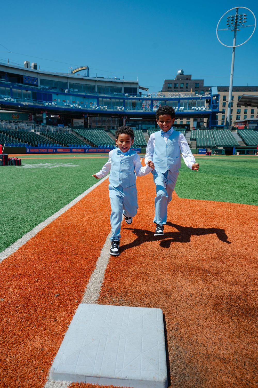 Two young boys in matching light blue vests and pants run toward a white base on a baseball field on a sunny day.