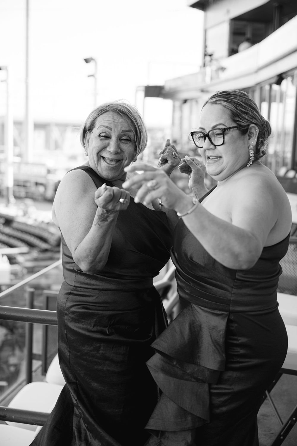 Two smiling women in dark satin dresses dance joyfully together on an outdoor terrace in a black and white celebration photo.