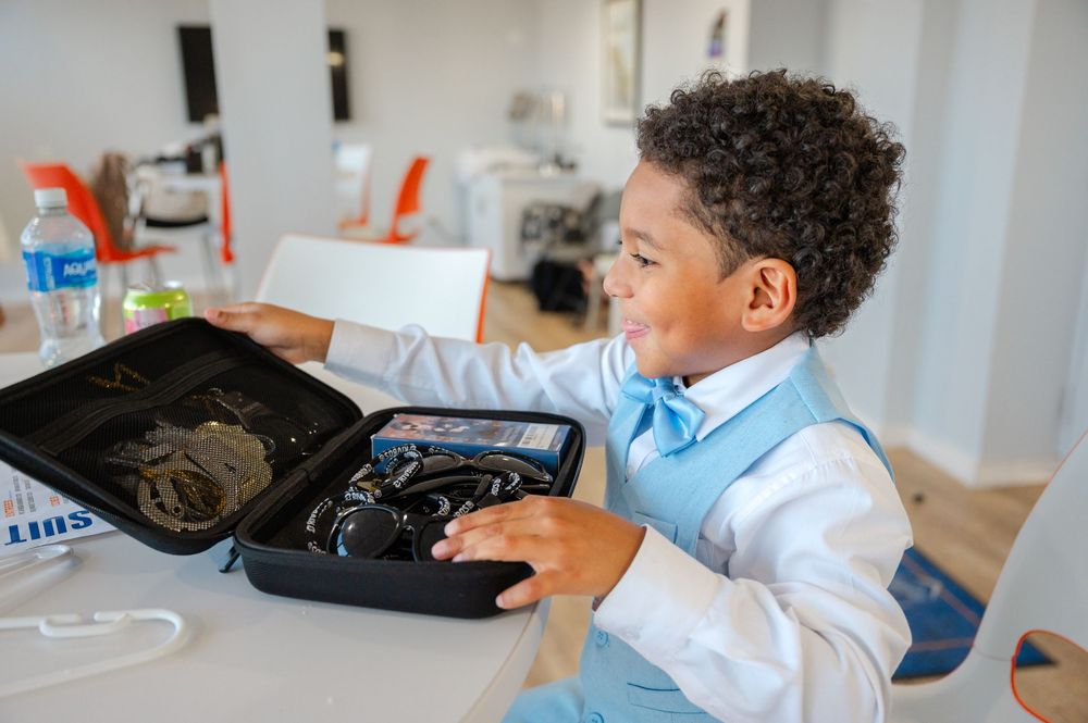 Smiling boy in a light blue vest and bow tie opens a black carrying case filled with headphones and audio equipment at a table.