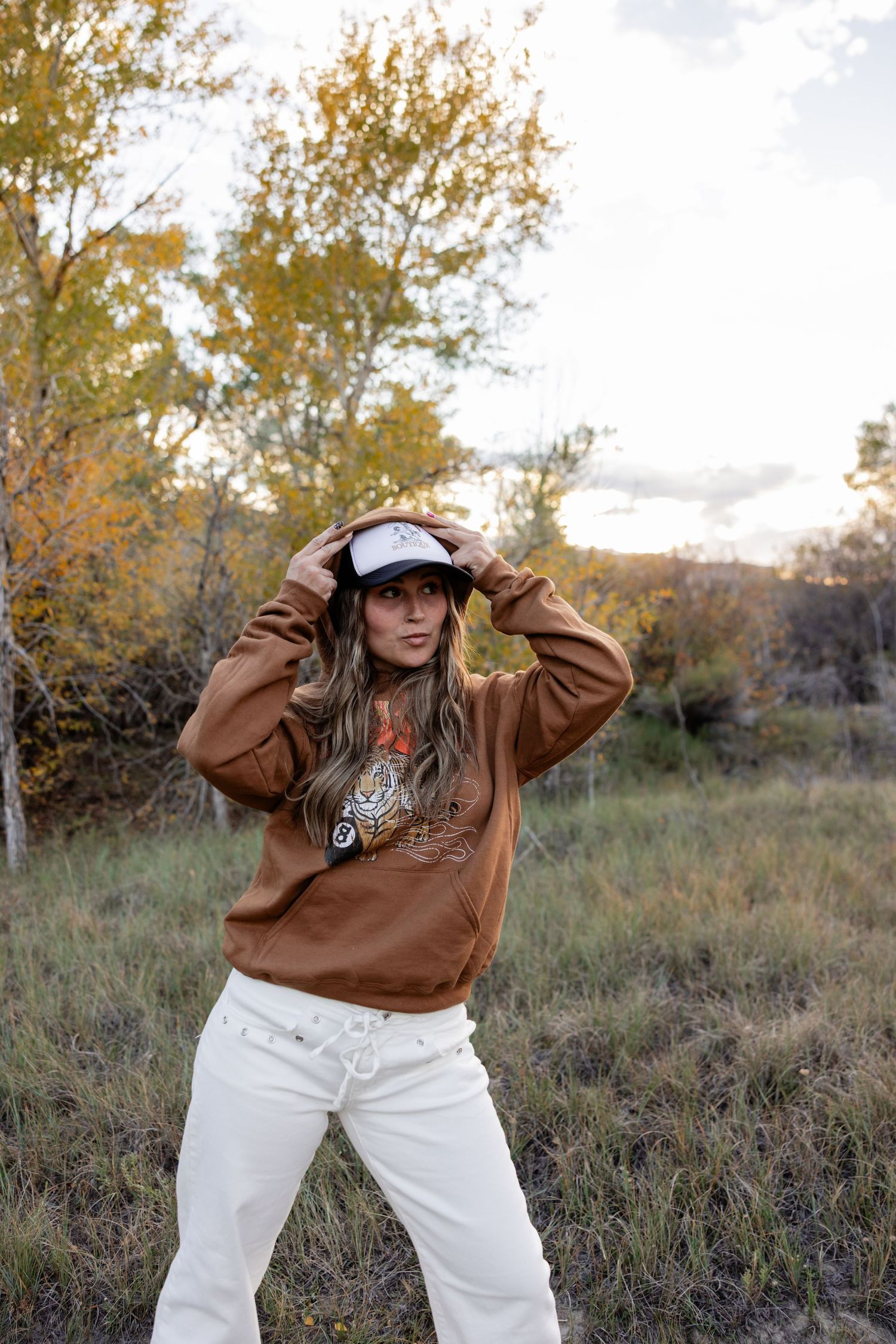 Woman in a brown graphic hoodie, cream pants and a trucker hat poses in a grassy field with golden autumn trees behind her.