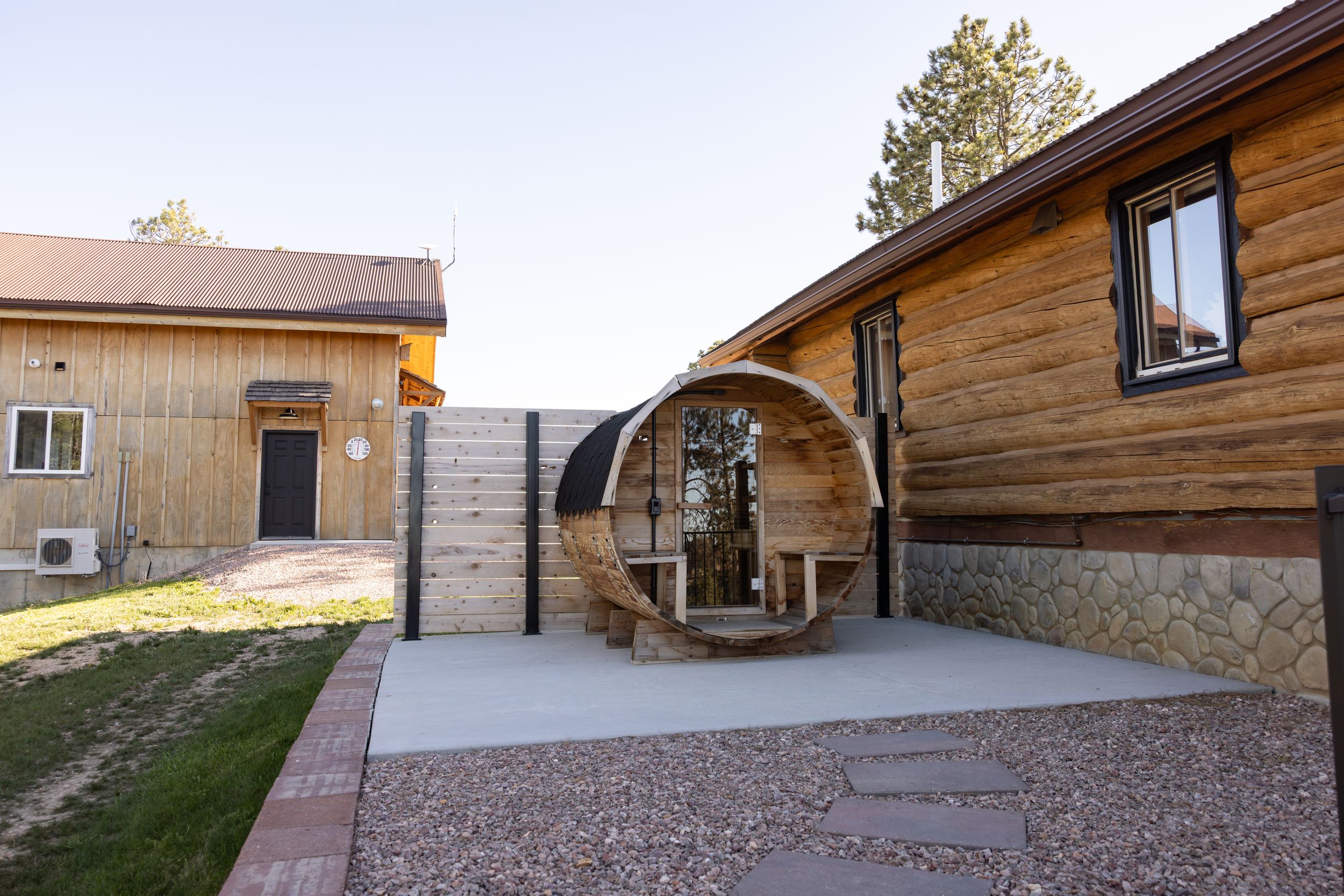 Barrel sauna on a concrete patio beside a rustic log cabin with stone foundation and a wooden outbuilding under a clear blue sky.