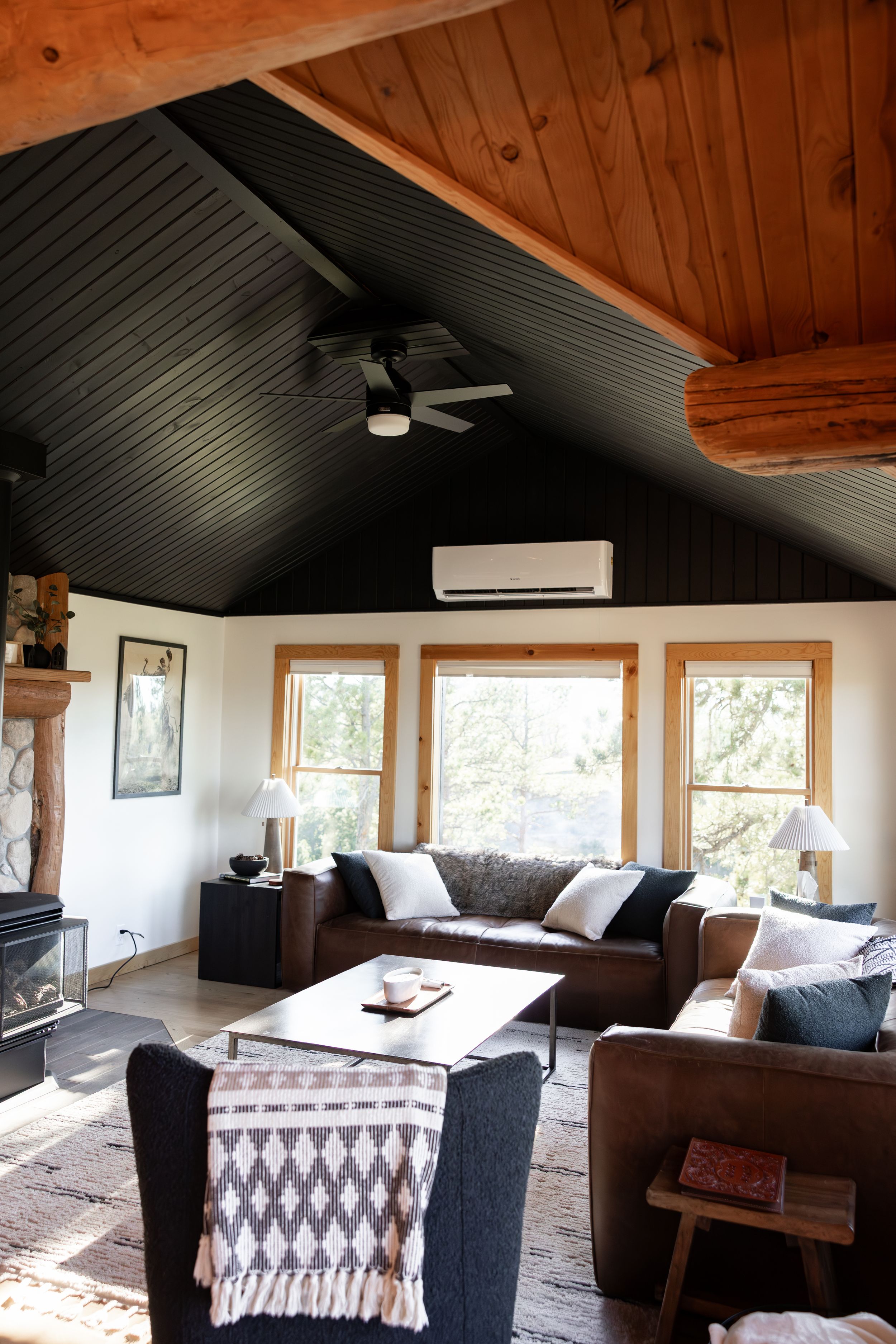 Cozy cabin living room with dark vaulted ceiling, exposed log beams, leather sofas, large windows, and a wood stove.
