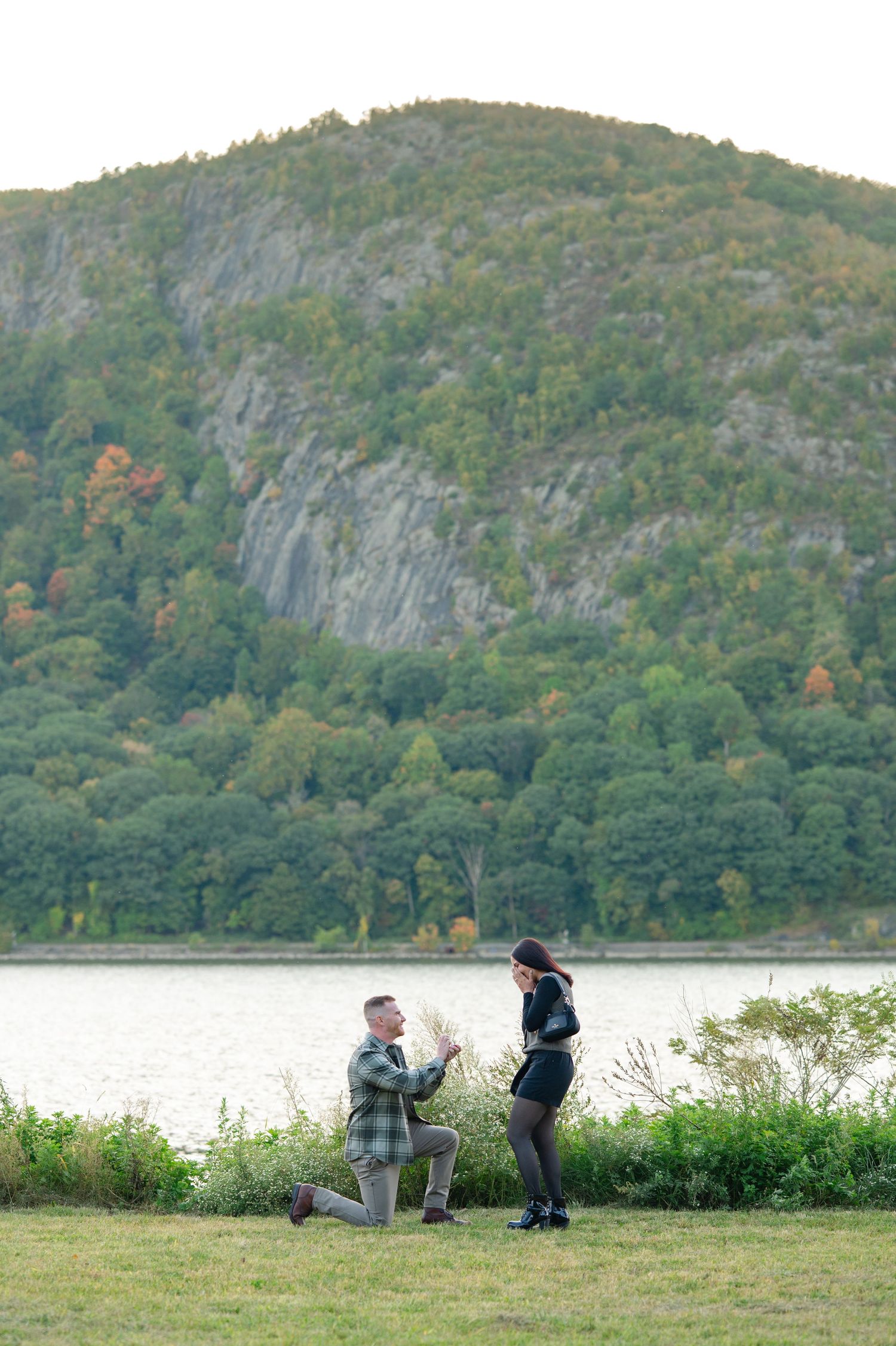 A man kneels on one knee proposing to a woman in Dockside Park next to the Hudson River in Cold Spring during a Hudson Valley proposal.