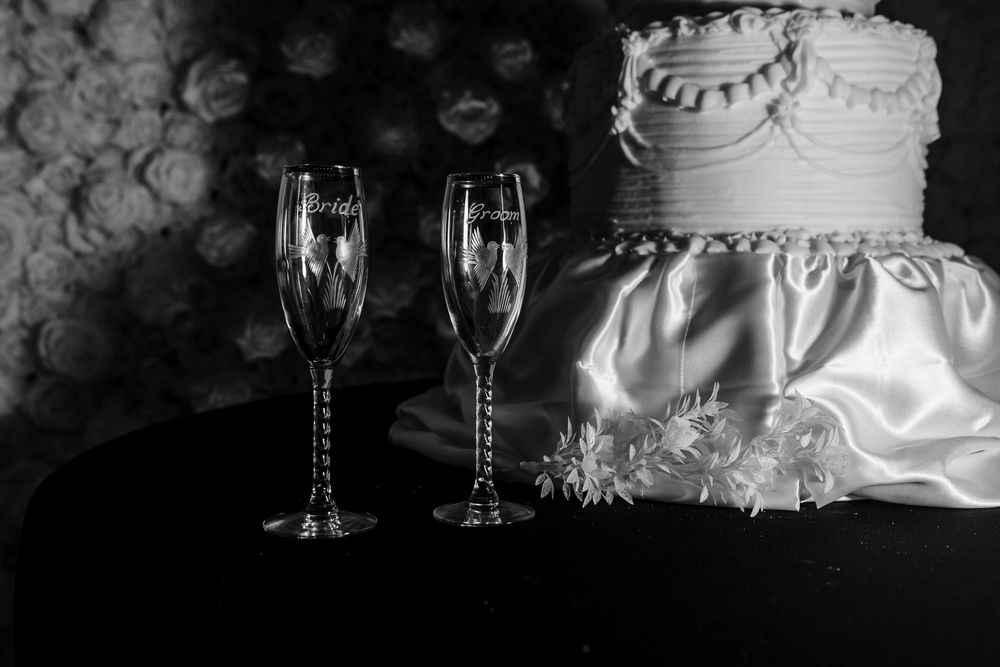 Black and white photo of two engraved bride and groom champagne flutes beside a tiered wedding cake draped in satin with flowers.