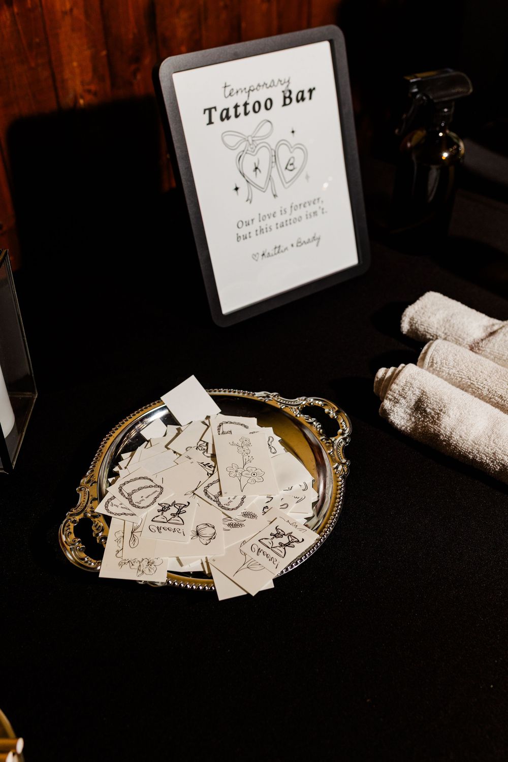 Wedding temporary tattoo bar sign beside a silver tray of tattoo sheets and rolled white towels on a black table.