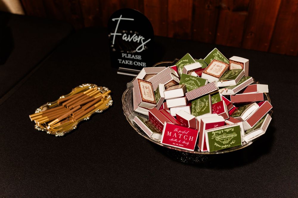 Wedding favors table with a silver tray of red and green personalized matchboxes beside a tray of gold sticks and a Favors sign.