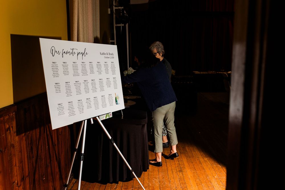 Woman in a navy top and gray pants stands beside a wedding seating chart on an easel in a warmly lit wooden-floored venue.