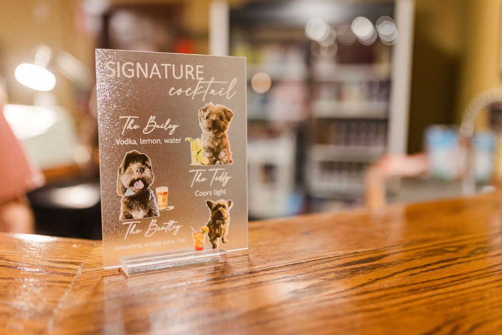 Acrylic Signature Cocktail menu stand on a wooden bar featuring photos of three dogs beside drink names and ingredients.