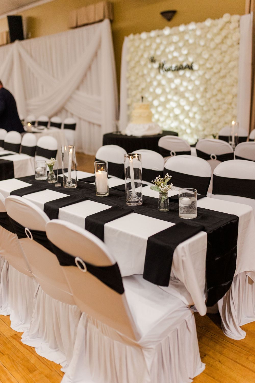 Elegant wedding reception table with black runners, white chair covers, candles and flowers, in front of a white floral backdrop and cake.