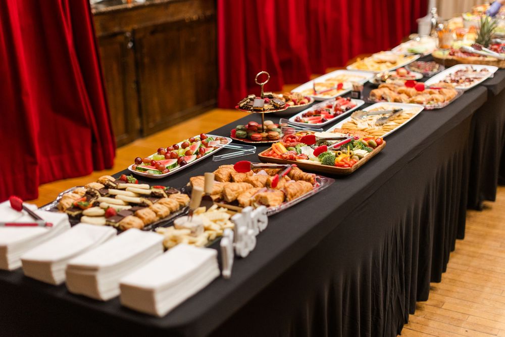 Buffet table with black linen displaying platters of assorted canapés and appetizers, with stacked white napkins and red curtains behind.