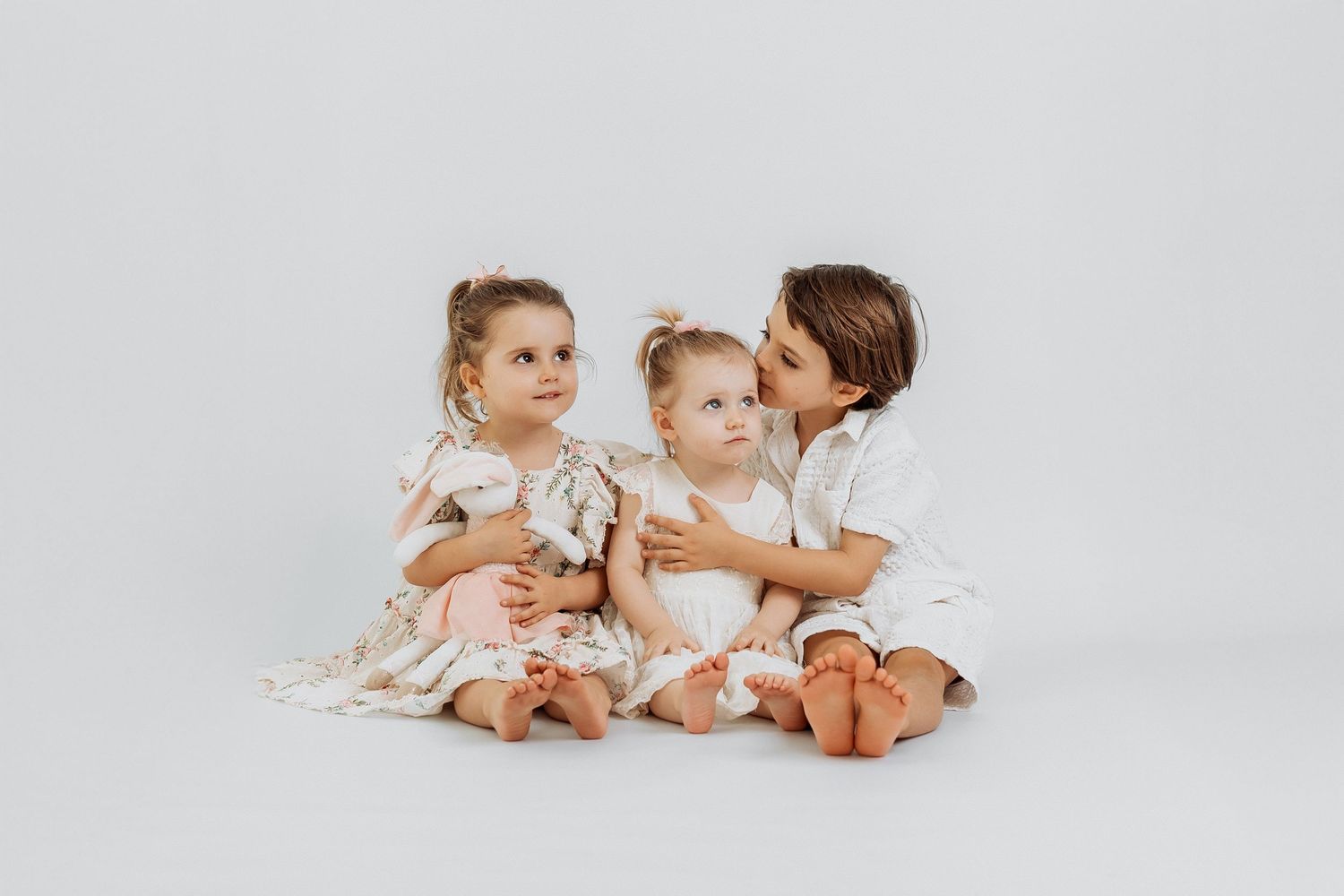 Three young siblings sitting together and hugging on a white backdrop during a studio family photoshoot in Dubai