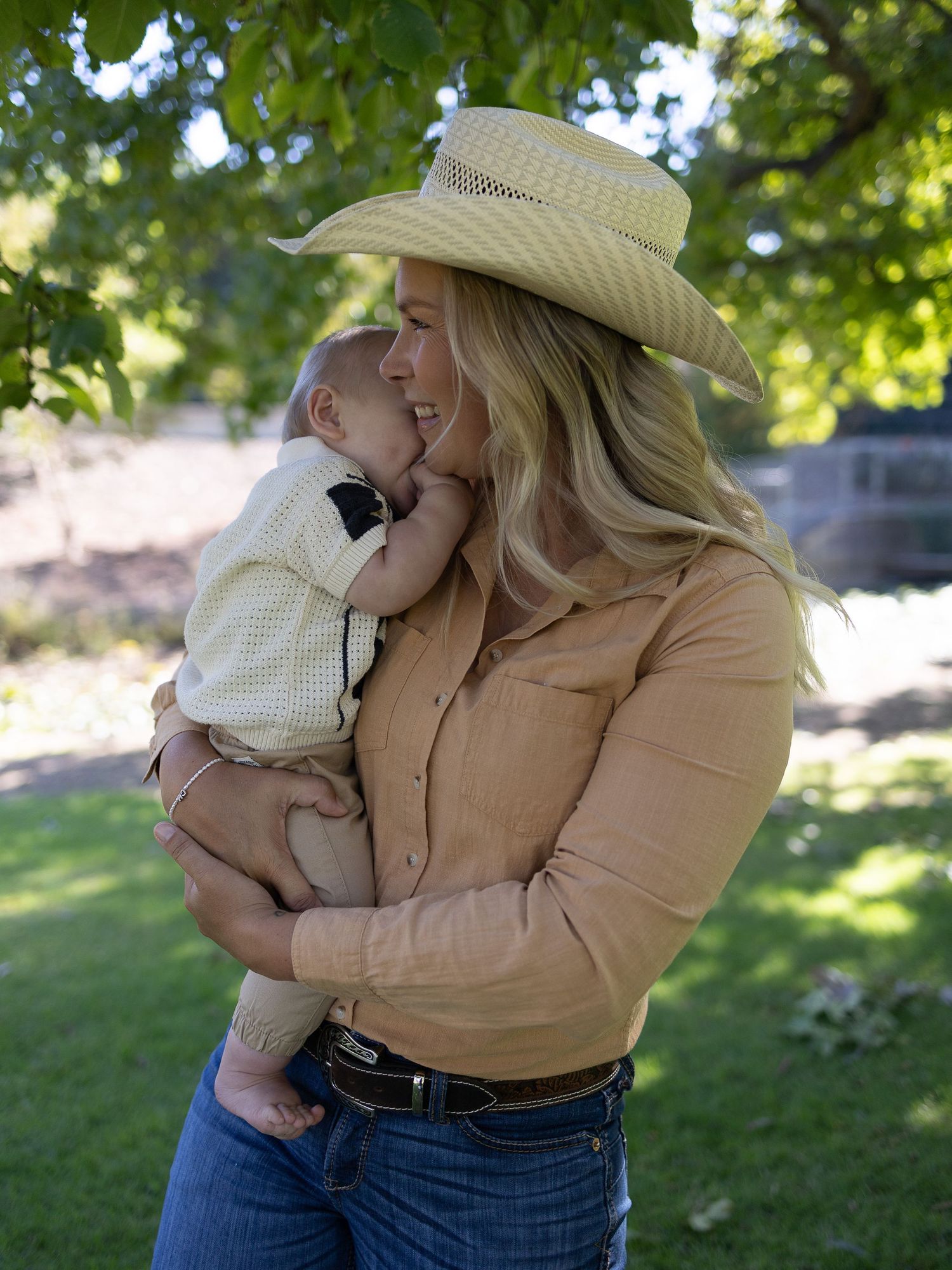 Smiling woman in a straw cowboy hat and tan button-up shirt holds a baby close outdoors on a sunny lawn beneath leafy green trees.