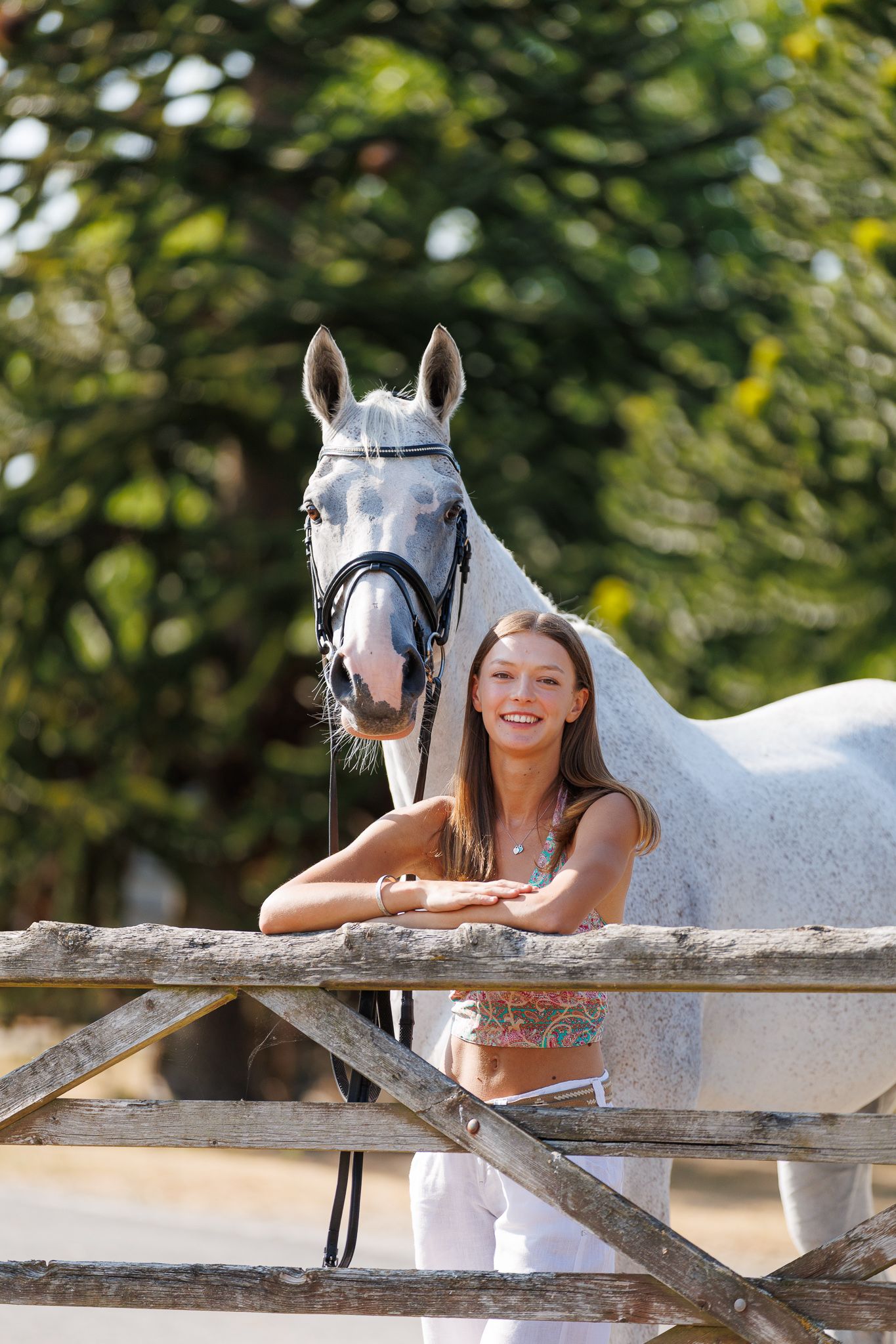 Woman in light blue gown walking with a bay horse on a sunlit tree-lined gravel path.