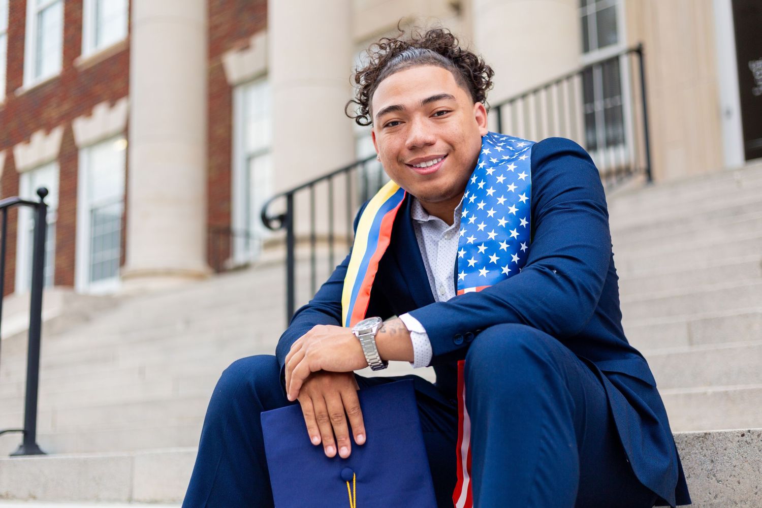 Graduate in a navy suit and colorful stole poses on brick building steps with white columns and a black metal railing.