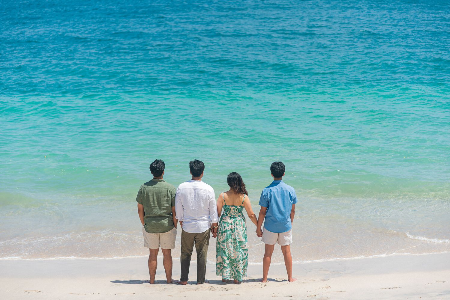 Four people stand with backs to camera on a white sandy beach, gazing at turquoise ocean waters on a sunny day.
