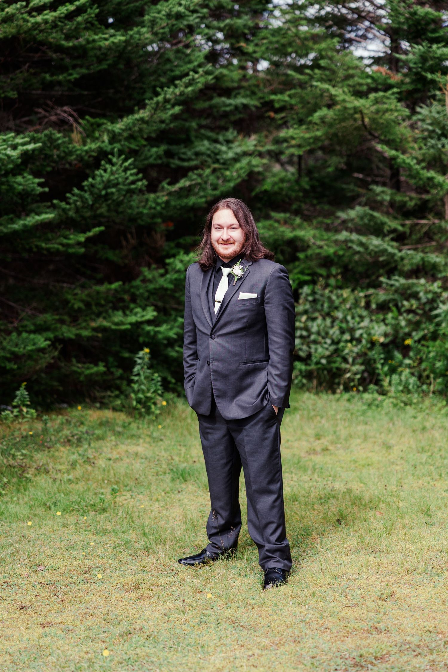 A groom in a dark gray suit with a floral boutonniere stands smiling on grass with evergreen trees behind him.