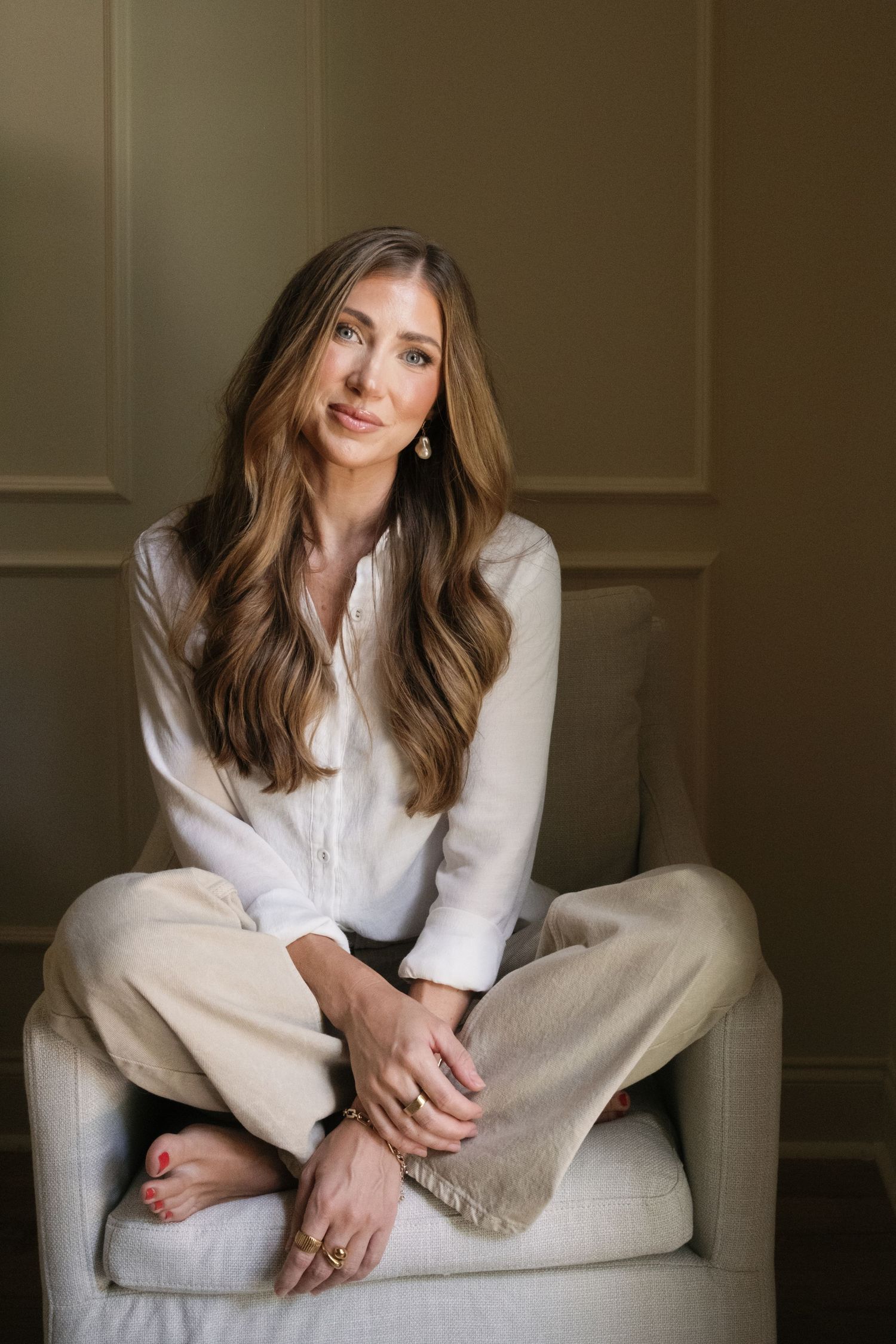 Woman with long brown hair sitting cross-legged in a white blouse and cream pants in a neutral room.
