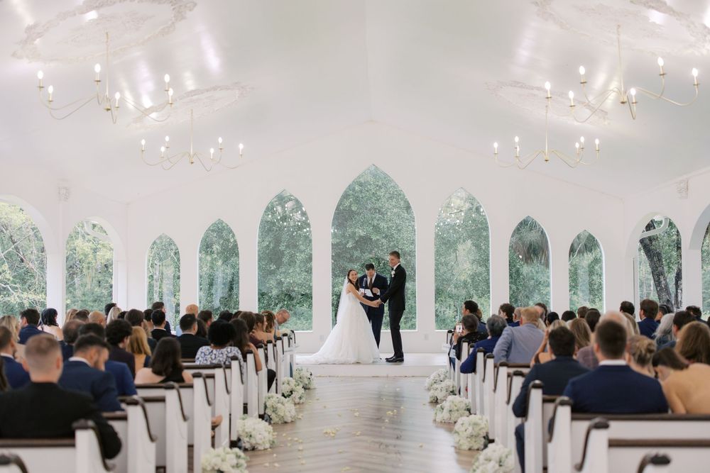 Elegant white chapel wedding ceremony with bride and groom at altar, arched windows, chandeliers, and seated guests on wooden pews.